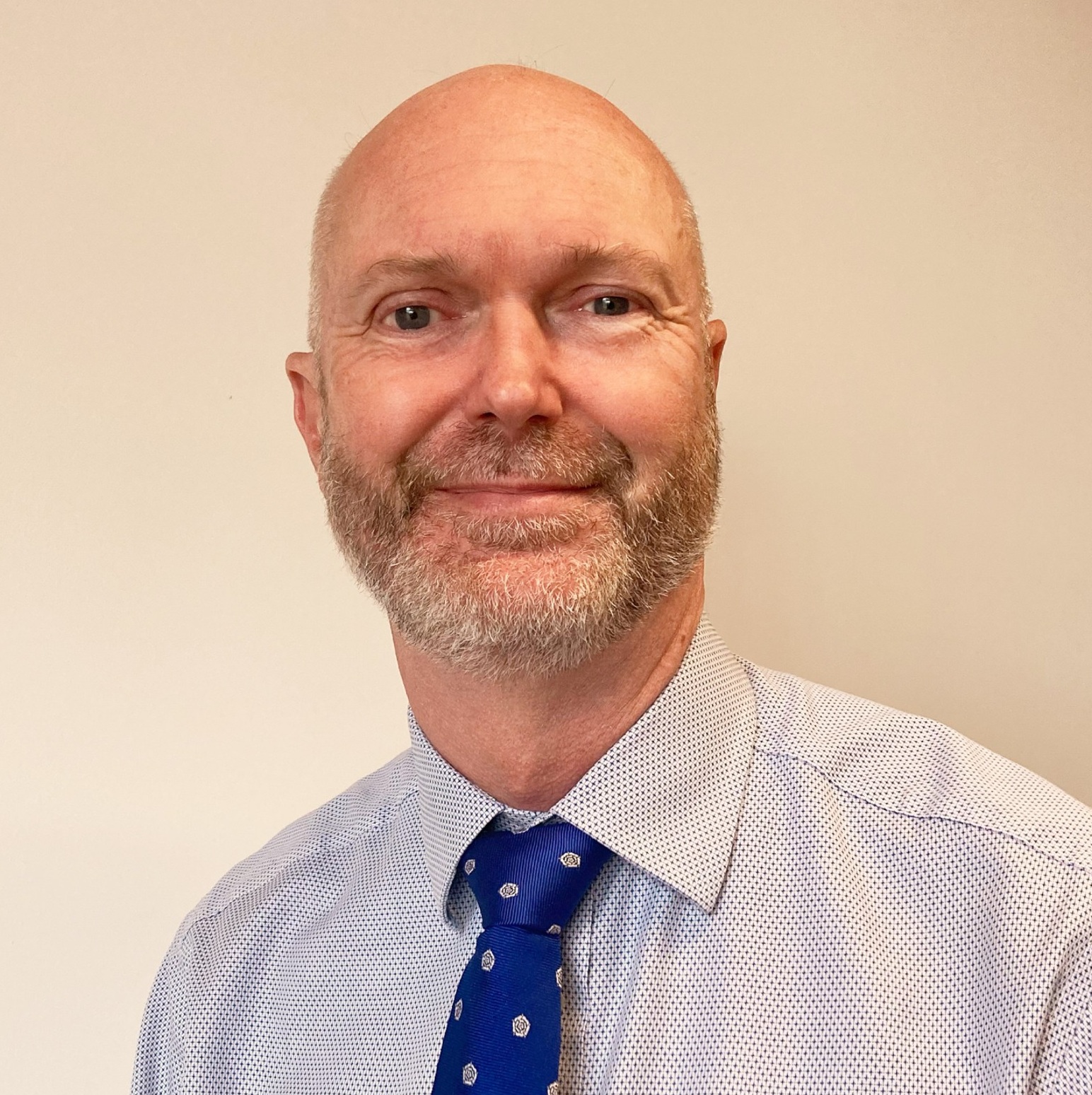 A middle-aged man with a bald head and trimmed gray beard, smiling softly. He is wearing a light blue shirt and a blue tie with small white spots, posing in front of a plain light-colored background.