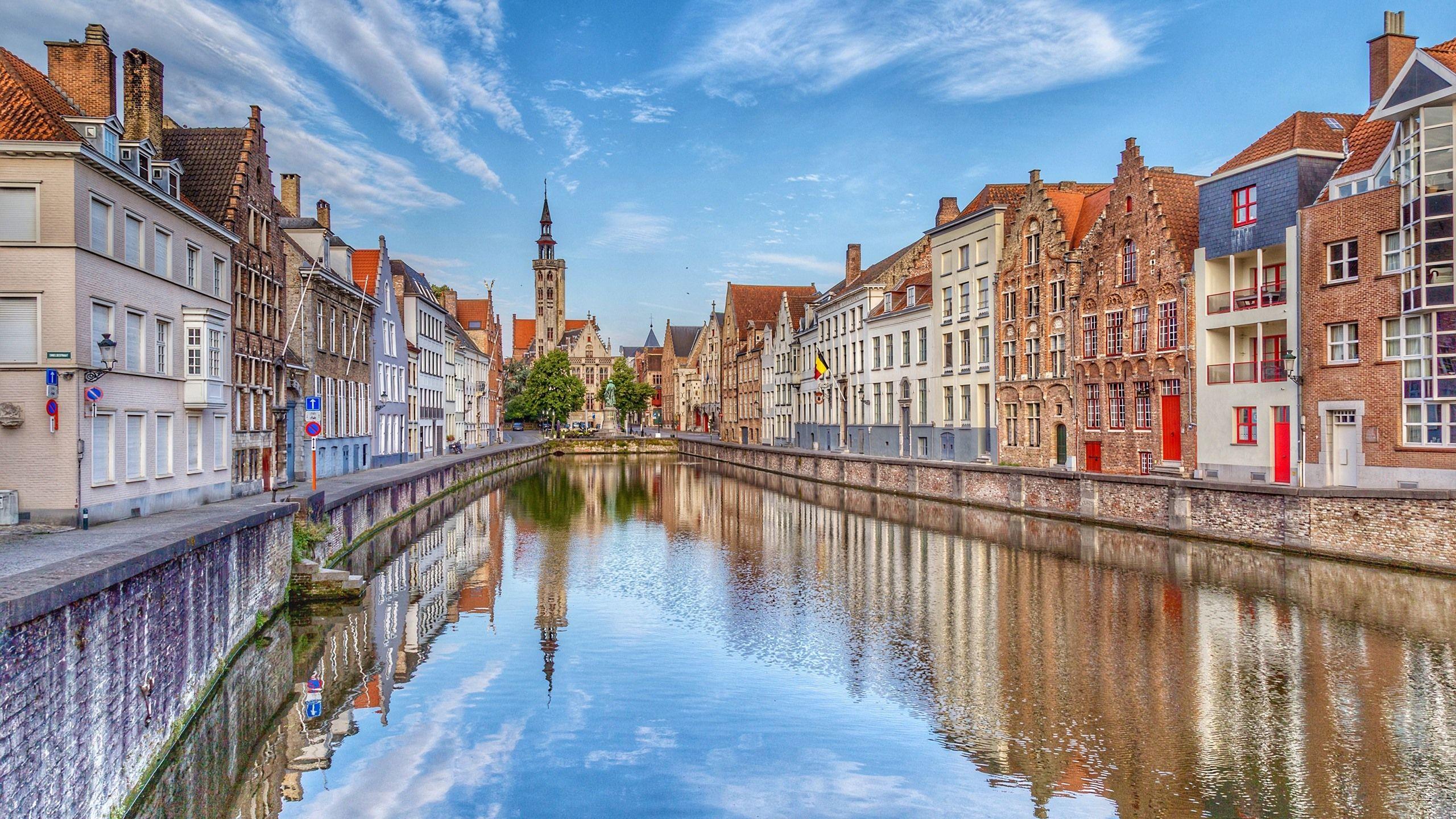 A canal lined with colorful historic buildings and a church tower in the background reflects in the calm water under a blue sky with wispy clouds.