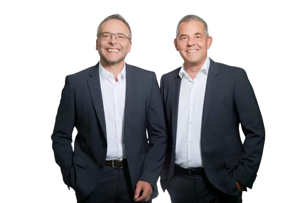 Zwei Männer in dunklen Anzügen und weißen Hemden stehen nebeneinander und lächeln in die Kamera, vor einem schlichten weißen Hintergrund.