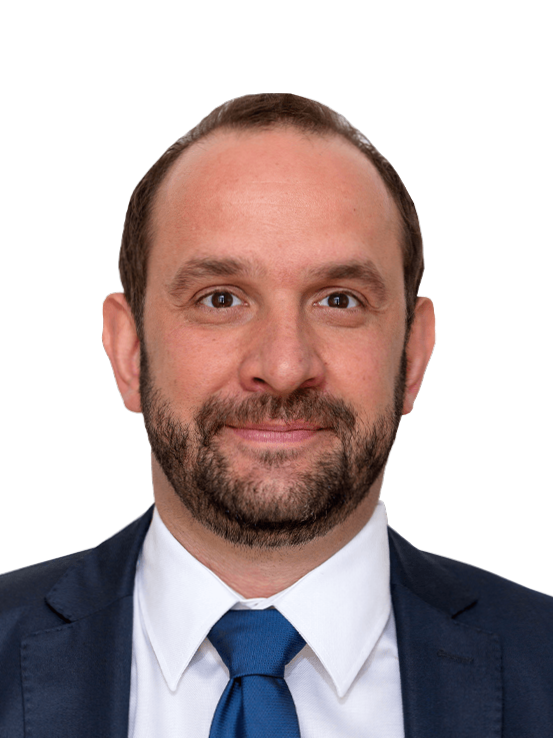 Ein Mann mit kurzen braunen Haaren und Bart, der einen blauen Anzug, ein weißes Hemd und eine blaue Krawatte trägt und vor einem schlichten Hintergrund in die Kamera lächelt.