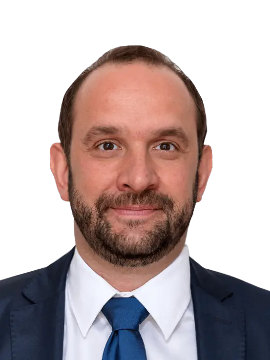 Ein Mann mit kurzen braunen Haaren und Bart, der einen blauen Anzug, ein weißes Hemd und eine blaue Krawatte trägt und vor einem schlichten Hintergrund in die Kamera lächelt.