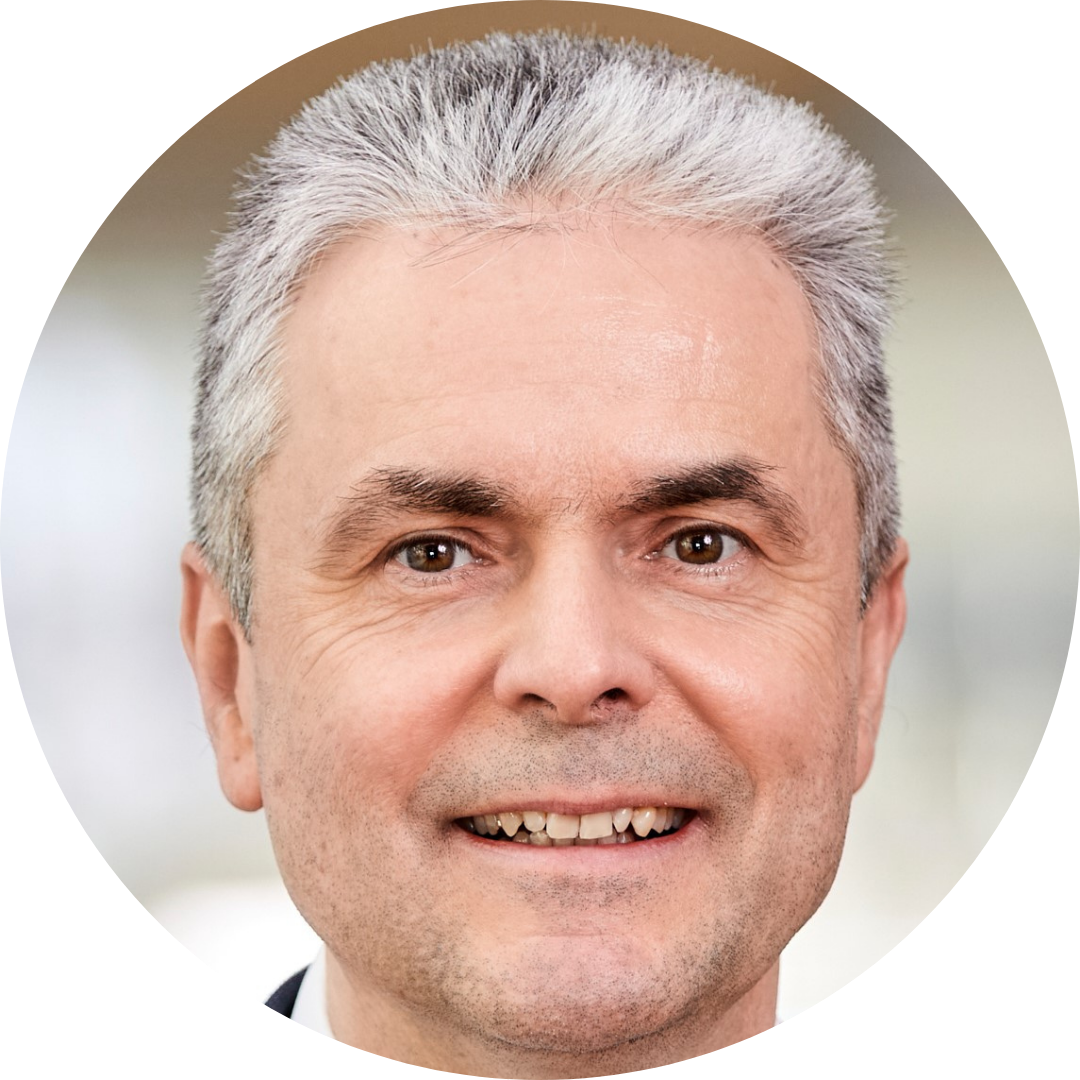 A middle-aged man with short, gray hair smiles at the camera. He has light skin, brown eyes, and is photographed against a softly blurred background at the BCM Sundown Symposium Frankfurt.