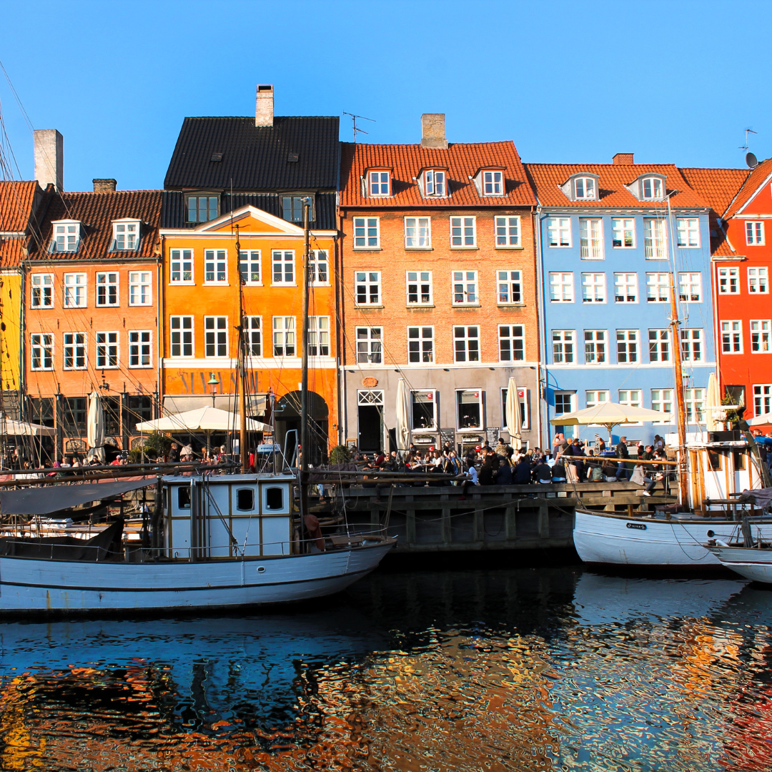 Colorful row houses line a waterfront in Nyhavn, Copenhagen, Denmark, with boats docked in the canal and people sitting at outdoor cafes under a clear blue sky.