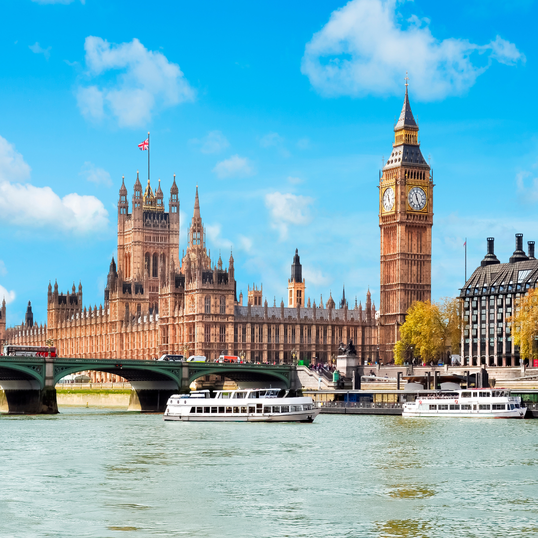 The Houses of Parliament and Big Ben in London, with boats on the River Thames and Westminster Bridge in the foreground under a bright blue sky.