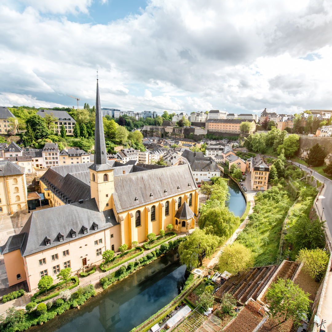 Aerial view of a historic European city with a large yellow church, a tall spire, a river, lush greenery, and surrounding traditional buildings under a partly cloudy sky.