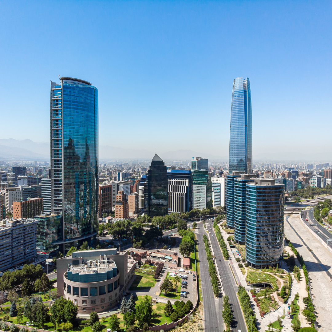 Aerial view of modern skyscrapers and office buildings in Santiago, Chile, with clear skies and mountains faintly visible in the background. A wide road runs through the cityscape with green areas on both sides.