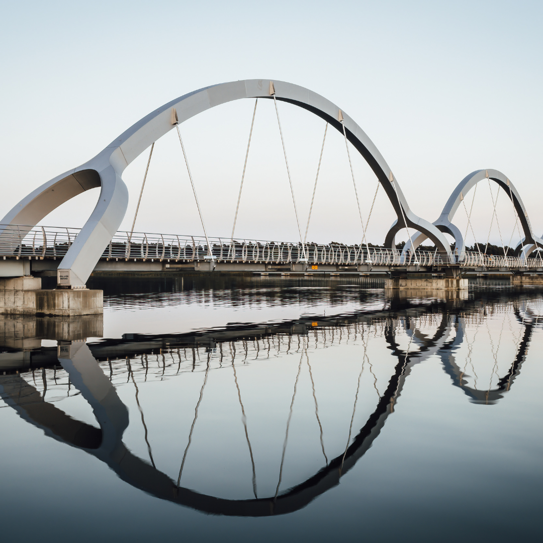 Modern arched pedestrian bridge with cables, reflected symmetrically in calm water below, under a clear sky.