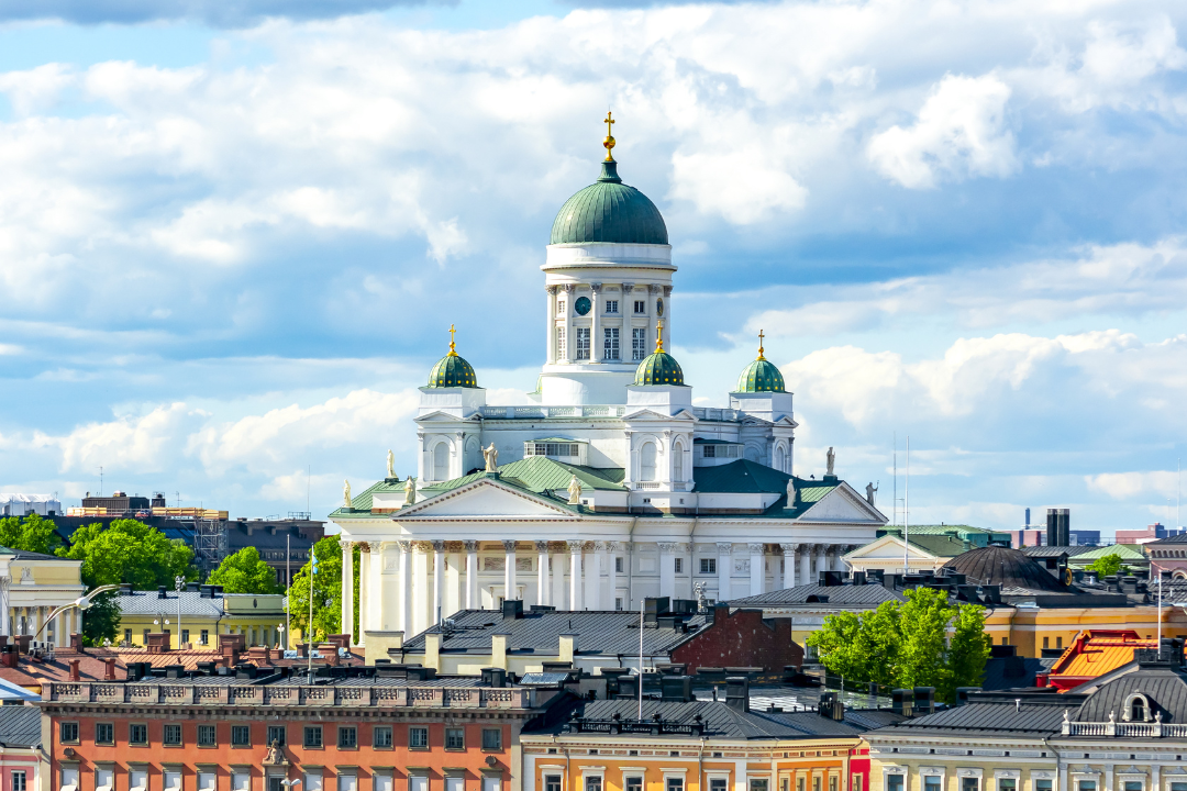 Eine Ansicht von Helsinki am Wasser mit farbenfrohen historischen Gebäuden, belebten Straßen und dem weißen Dom von Helsinki mit seinen grünen Kuppeln, die sich im Hintergrund unter einem teilweise bewölkten Himmel erheben.