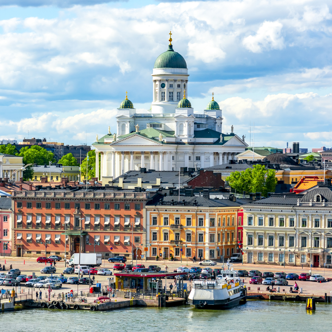 View of Helsinki Cathedral with its green domes rising above colorful historic buildings, cars, and a docked boat by the waterfront under a partly cloudy sky.