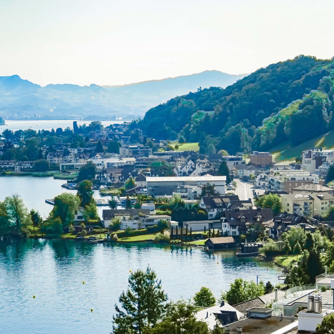 A picturesque lakeside town with houses and buildings surrounded by lush green hills, with mountains in the background and a calm blue lake in the foreground on a sunny day.