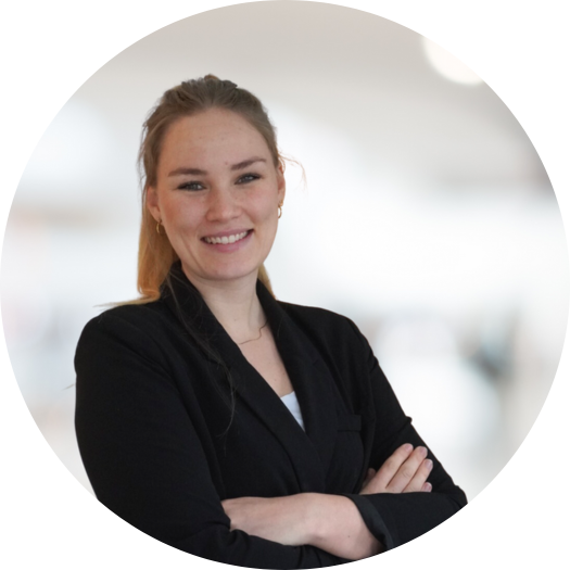 A woman with light brown hair tied back, wearing a black blazer and white top, stands with arms crossed and smiles confidently at the camera, ready to guide your Experience Tour Madrid. The background is blurred and light-colored.