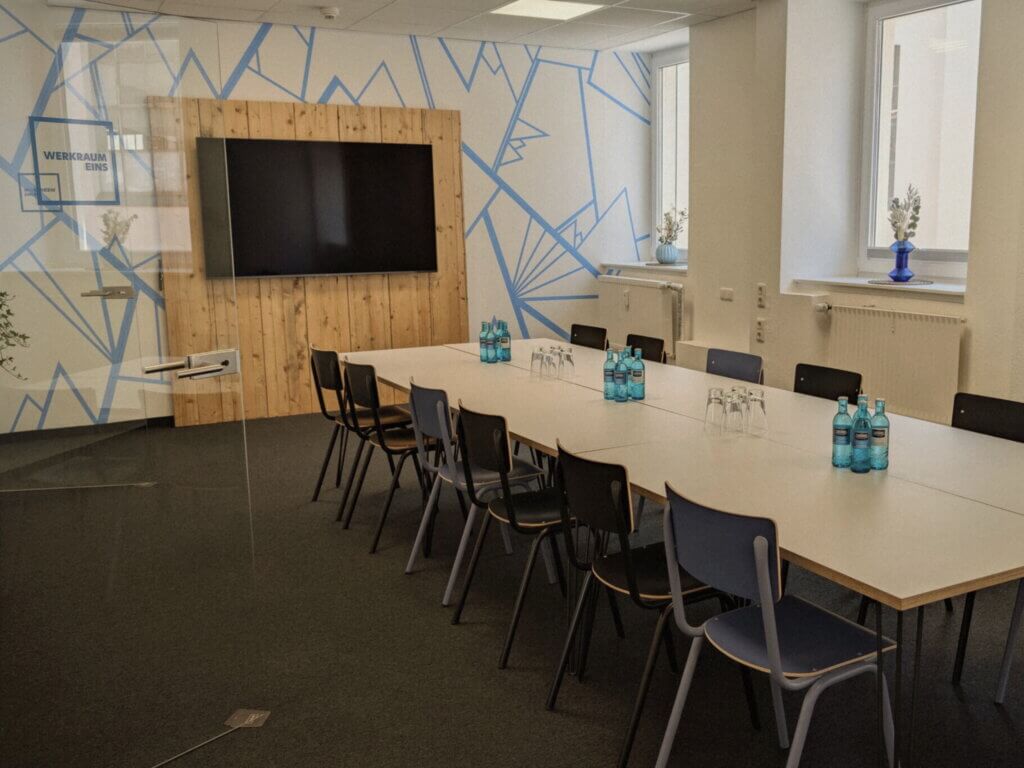 A modern conference room with a long white table, black chairs, a large TV screen on a wooden panel, water bottles, glasses, and decorative vases on the window sills. Blue geometric patterns are on one wall.