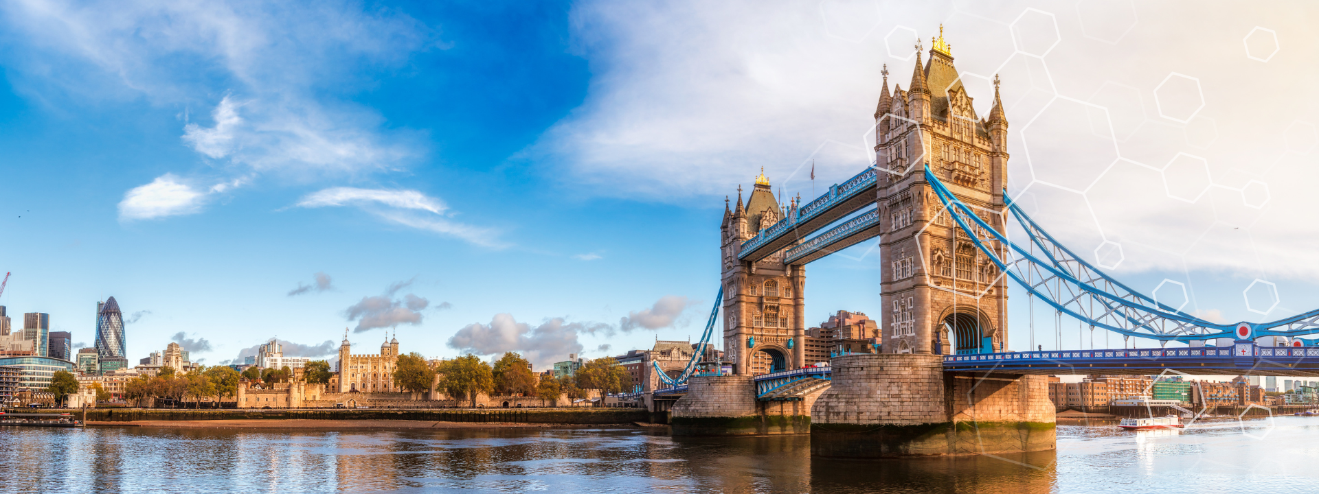 Panoramic view of London’s Tower Bridge over the River Thames on a bright day, with modern and historic buildings visible along the riverbank under a blue sky with scattered clouds.