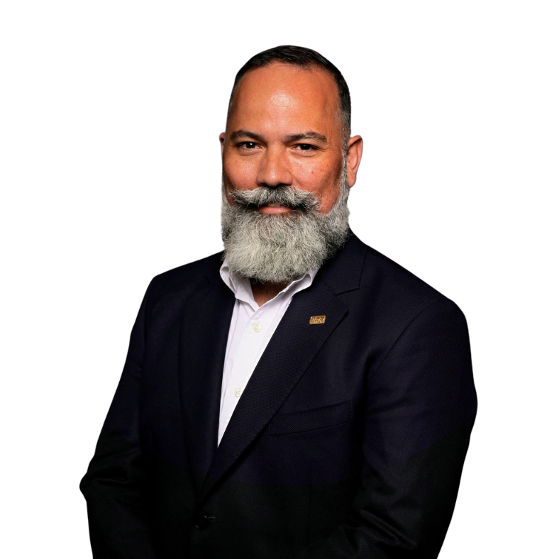 A man with a thick gray beard and mustache, wearing a dark suit jacket over a white shirt, poses against a plain white background, smiling slightly with arms relaxed by his sides, exuding calm confidence even in an emergencia.