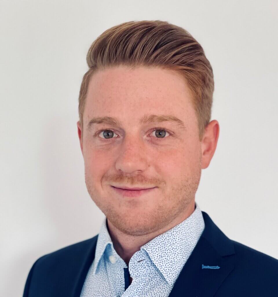A young man with short, neatly styled light brown hair, wearing a navy blazer and a white patterned shirt, smiles slightly while standing in front of a plain white background.