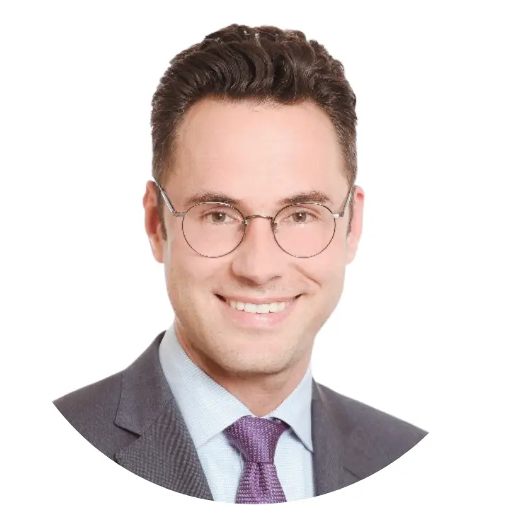 A man with short, dark hair wearing round glasses, a grey suit, a light blue shirt, and a purple tie smiles at the camera. The background is white—highlighting confidence like eine starke Schnittstelle für den Ernstfall: FACT24 ENS+ in Winguard.