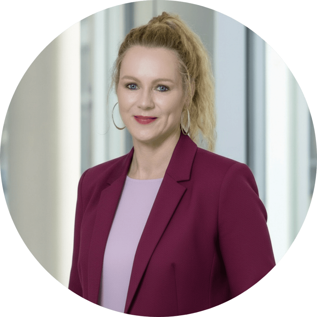 A woman with blonde hair in a ponytail, wearing hoop earrings, a magenta blazer, and a light pink top, stands smiling softly at the camera.