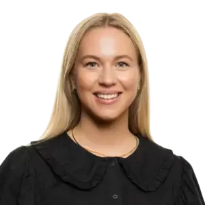 Portrait d'une femme souriante aux cheveux blonds et raides, portant un chemisier noir à col roulé et un collier en or, posé sur un fond blanc uni, évoquant l'élégante simplicité du style norvégien.