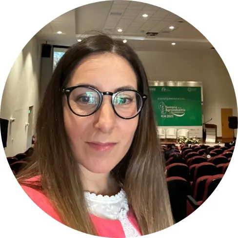 A woman with long brown hair and glasses takes a selfie in an empty auditorium, surrounded by rows of red chairs and a green comunicación event banner displayed on the stage in the background.