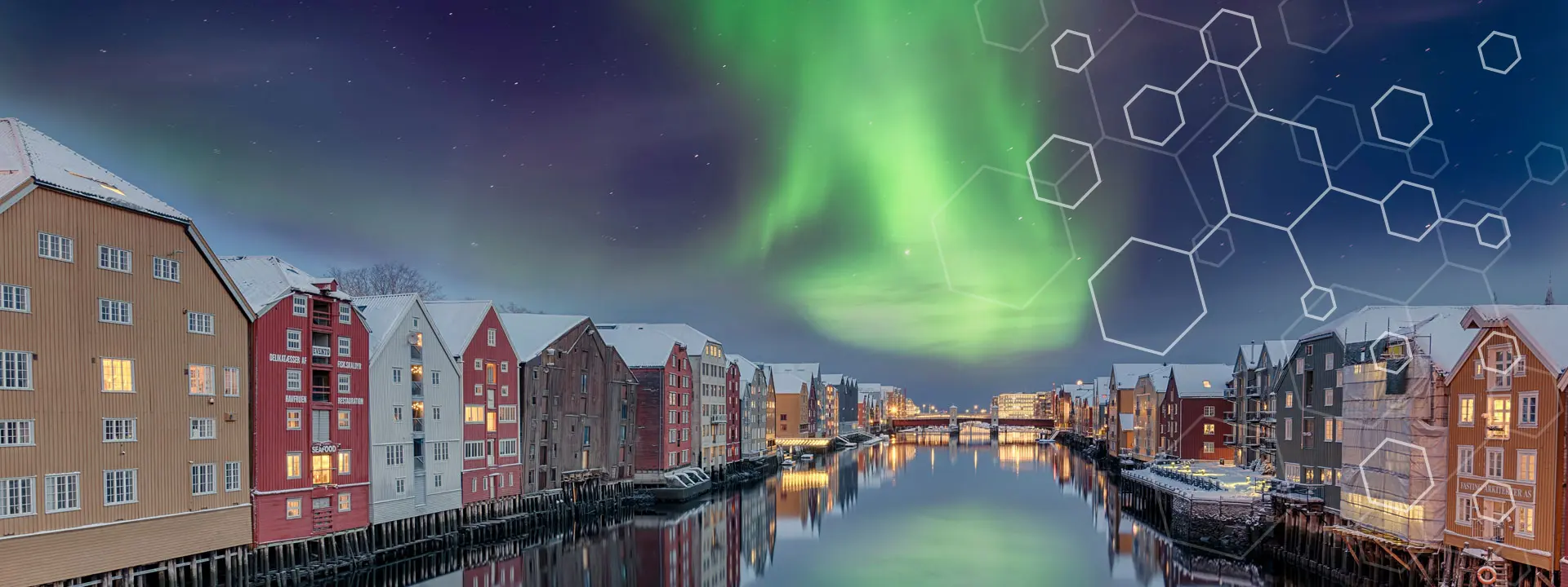 Colorful buildings in Norway line a calm river under the northern lights at night, with geometric hexagon patterns overlaid on the sky. Snow covers the rooftops and shimmering reflections dance in the water.