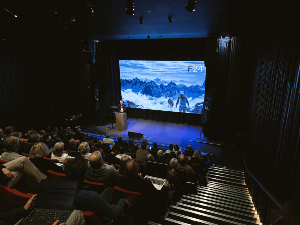 A speaker presents on stage in a dark auditorium to an audience, with a large screen behind showing a snowy mountain scene labeled "F24"—part of the Experience Tour on-demand.