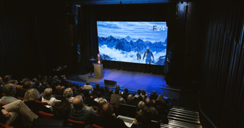 A speaker presents on stage in a dark auditorium to an audience, with a large screen behind showing a snowy mountain scene labeled "F24"—part of the Experience Tour on-demand.