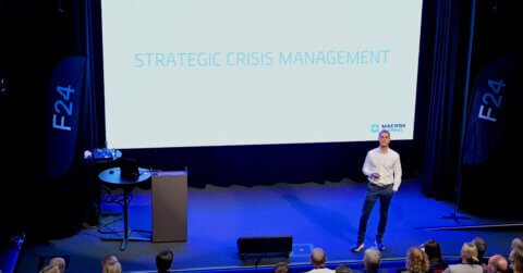 A speaker stands on stage in front of a large screen displaying the words “Strategic Crisis Management” in a dark auditorium, addressing an audience seated below.