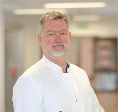 A middle-aged man with short gray hair and a beard, wearing a white lab coat, stands in a bright hospital setting, offering Einblicke aus dem Klinikum Oldenburg zu Krisenmanagement und Alarmierung im Klinikalltag.
