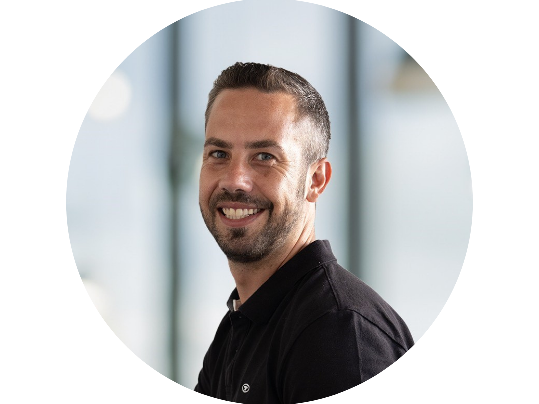A man with short dark hair and a beard, wearing a black shirt, smiles at the camera against a blurred background with large windows—capturing the spirit of Krisenmanagement und Alarmierung im Klinikalltag im Klinikum Oldenburg.