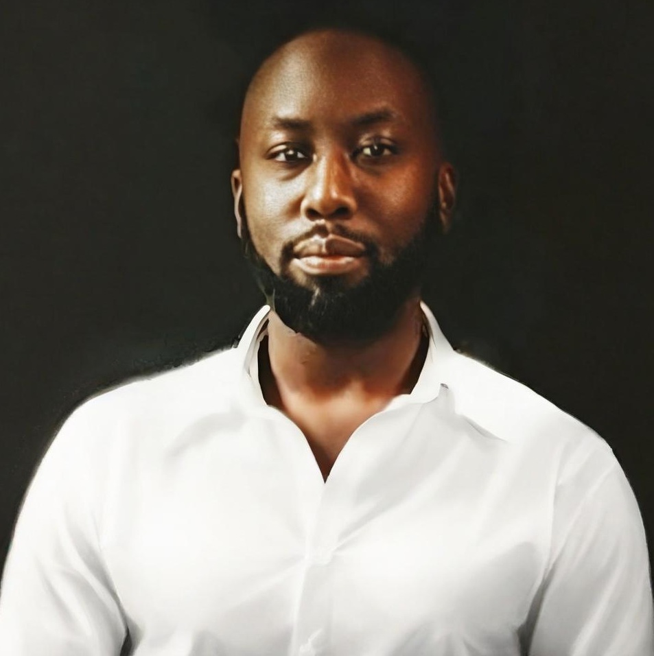 A man with a beard and mustache wearing a white button-up shirt poses against a dark background, representing Petrofac during the F24 ENS+ Webinar. He has a neutral expression and short hair.