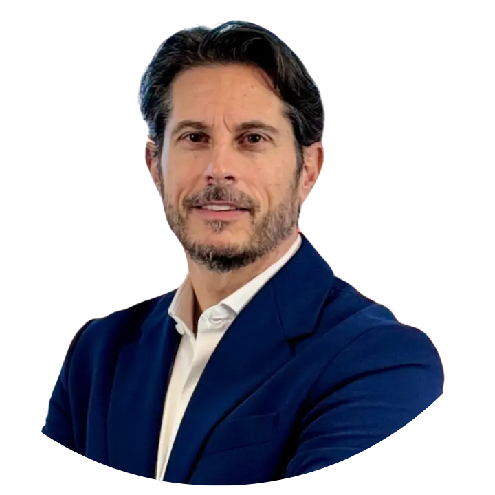 A man with dark hair and a beard, wearing a navy blue suit jacket and white shirt, poses KURZ UND BÜNDIG with arms crossed against a plain light background.