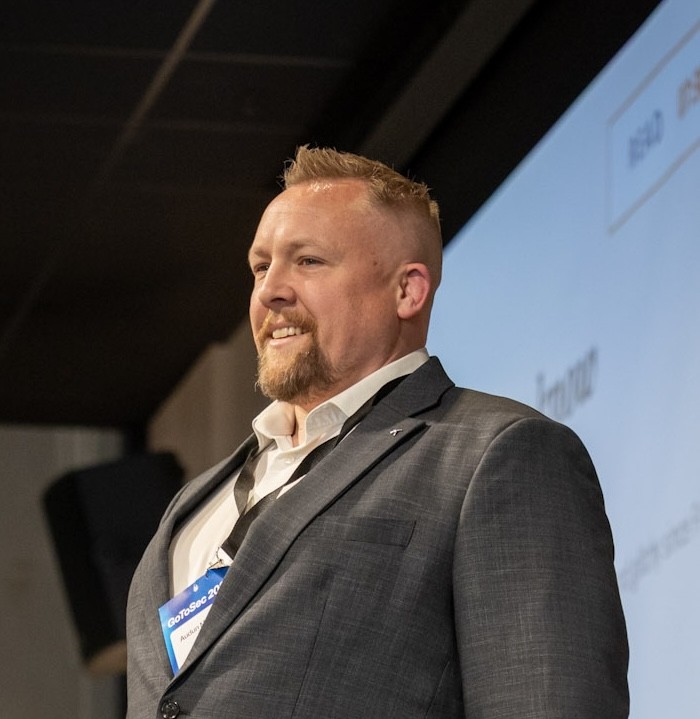 A man with short blond hair and a beard, wearing a gray suit and white shirt, stands and smiles in front of a presentation screen at a professional event.