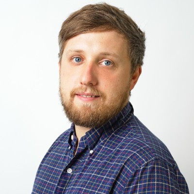 A man with light skin, brown hair, and a beard, wearing a blue and green checkered shirt, is facing slightly to the side and looking at the camera against a plain white background—ideal for promoting your upcoming F24 ENS+ Webinar.