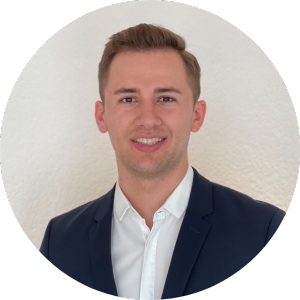 A young man with short light brown hair, wearing a white shirt and a dark blazer, smiles at the camera against a plain, cloud-white background.