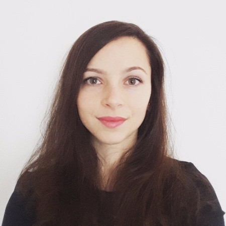 A young woman with long brown hair and fair skin looks directly at the camera, smiling slightly. She is wearing a black top and stands against a plain white background, embodying la résilience de la nation amidst geopolitical tensions.