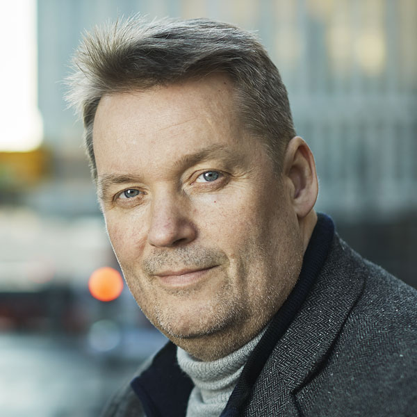 A middle-aged man with short, light brown hair and blue eyes, wearing a gray turtleneck and a dark patterned coat, stands outdoors in an urban setting—perhaps on his way to the "Webinar: Innføring i ROS-analyse" with blurred buildings in the background.