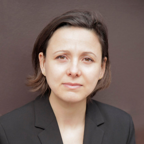 A woman with short brown hair wearing a black blazer looks at the camera with a neutral expression, standing against a plain brown background, evoking the theme of Résilience de la nation : Quand les tensions géopolitiques déstabilisent nos territoires.