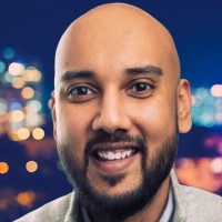 A smiling bald man with a beard and mustache, wearing a light-colored jacket, poses in front of a blurred cityscape with colorful lights at night, ready to host the F24 ENS+ Webinar for Petrofac.