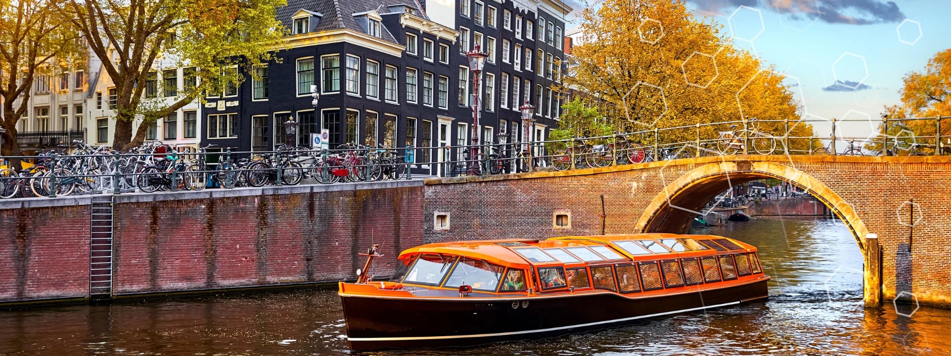 A glass-roofed canal boat passes under a brick bridge in Amsterdam, with historic buildings, trees with autumn leaves, and bicycles lining the canal at sunset. Subtle geometric patterns overlay the sky.