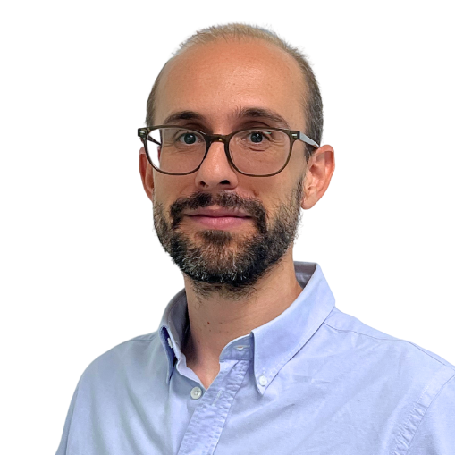 A man with short brown hair, a beard, and glasses, wearing a light blue button-up shirt, stands against a plain white background.