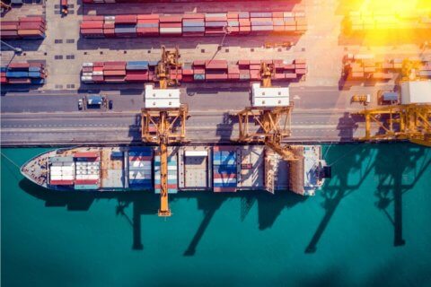 Flygfoto över ett lastfartyg som ligger dockat i en hamn, med kranar som hanterar containrar – nyckeln till Våra produkter: Varning och krishantering. Rader av färgglada fraktcontainrar kantar dockan bredvid det turkosa vattnet.