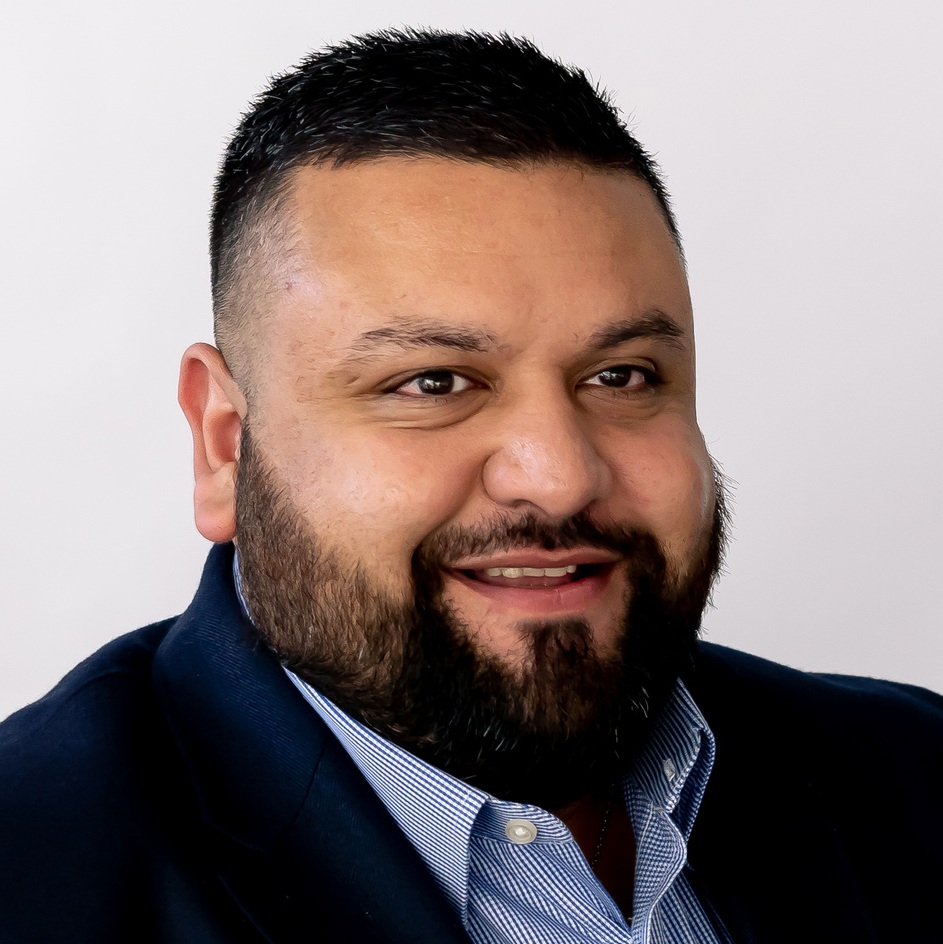 A man with short dark hair and a full beard, wearing a navy blazer and a light blue collared shirt, smiles while looking slightly to the side, ready to host the F24 ENS+ Webinar against a plain light background.