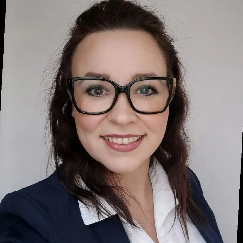 A woman with long dark hair, wearing black glasses, a navy blazer, and a white collared shirt, smiles at the camera against a plain light background, exuding confidence fit for the cloud technology sector.