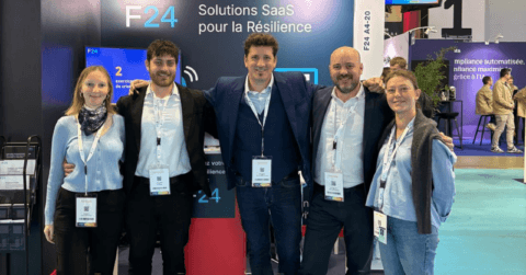 Five people stand smiling in front of an F24 booth at a conference, showcasing SaaS solutions for resilience. Retrouvez l'équipe F24 lors des salons 2026, wearing business attire and name badges amid brightly lit booths and attendees.