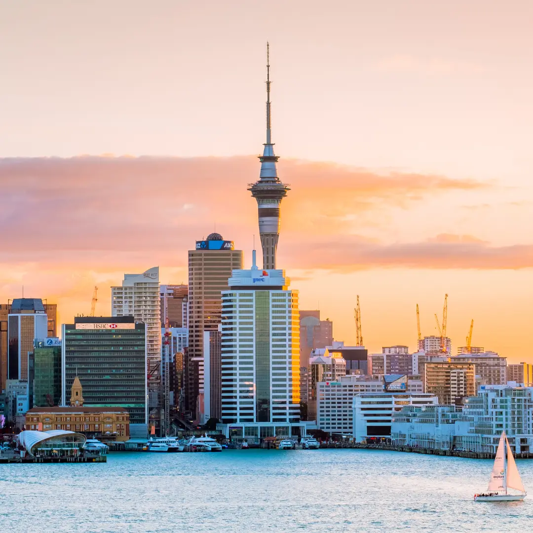 A city skyline at sunset with the tall Sky Tower in the center, surrounded by modern buildings. A sailboat glides on the water in the foreground as soft orange and pink hues fill the sky. For more details, contact f24.