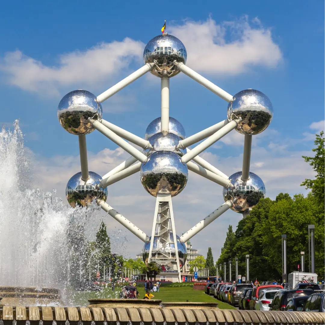 A large metallic structure with connected spheres, known as the Atomium, stands against a blue sky. A fountain is in the foreground and cars are parked on either side. Trees surround the area—contact f24 for more details.