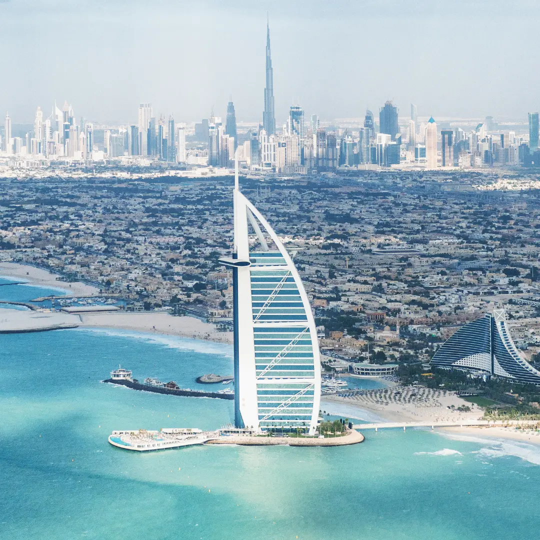 Aerial view of the sail-shaped Burj Al Arab hotel on its own island off the Dubai coastline, with turquoise water in the foreground and the city skyline, including Burj Khalifa, visible in the background—contact f24 for more details.