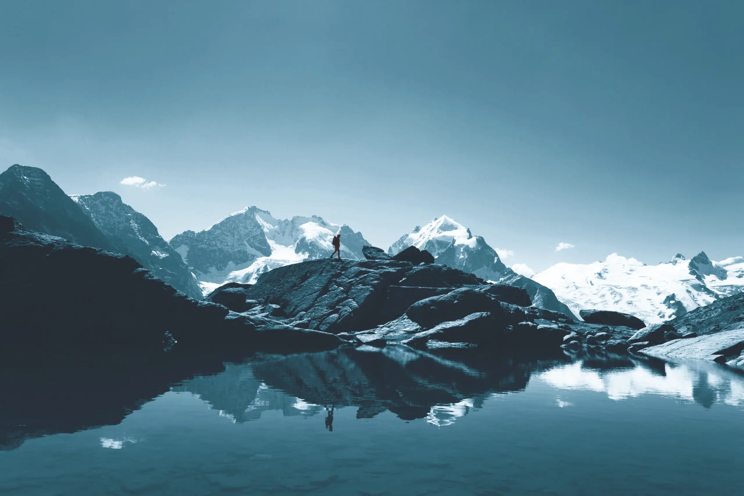Eine Person steht auf felsigem Gelände neben einem ruhigen Bergsee, im Hintergrund sind schneebedeckte Gipfel und ein klarer Himmel zu sehen. Die Szene spiegelt sich im stillen Wasser wider und schafft so eine friedliche und eindrucksvolle Landschaft.