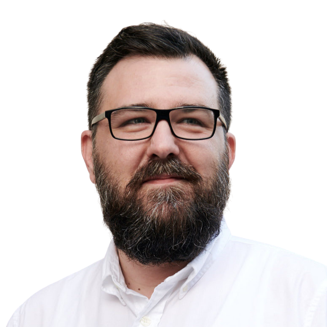 A man with dark hair, a full beard, and glasses is wearing a white collared shirt and looking slightly to the side, smiling softly against a plain, light background.