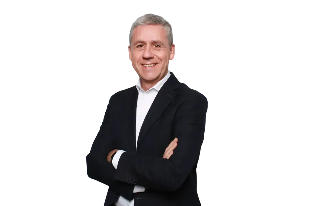 A smiling man with short gray hair stands with his arms crossed, wearing a dark suit jacket and a white dress shirt, against a plain white background.